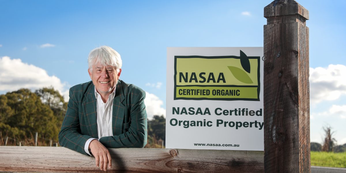 Board Member Mark Anderson stands at the gate of CRFT Wines next to a NASAA Certified Organic property sign. He is leaning against a fence post. There are blue skies.