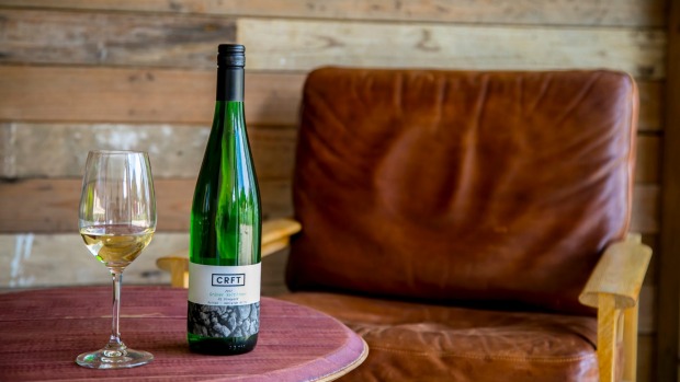 A bottle of organic CRFT wines with a half filled wine glass sits on the table with an old mid century chair next to it.